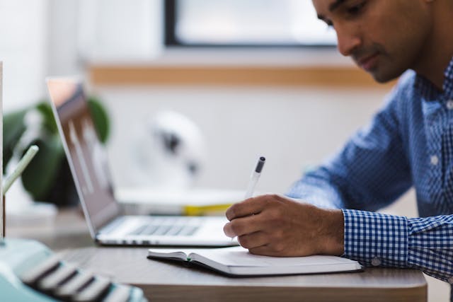 Person writing something down on a notepad with the laptop open in front of them